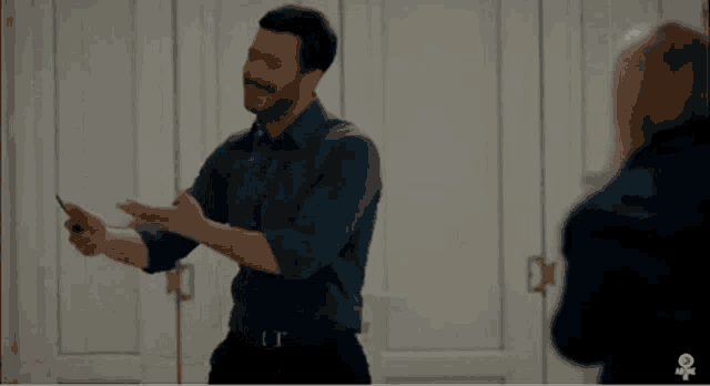 a man in a blue shirt is standing in front of a door holding a cell phone in his hand .