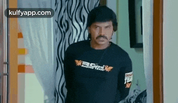 a man with a mustache is standing in a doorway wearing a black t-shirt and a necklace .