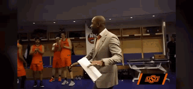 a man in a suit holds a piece of paper in front of a group of basketball players in orange jerseys