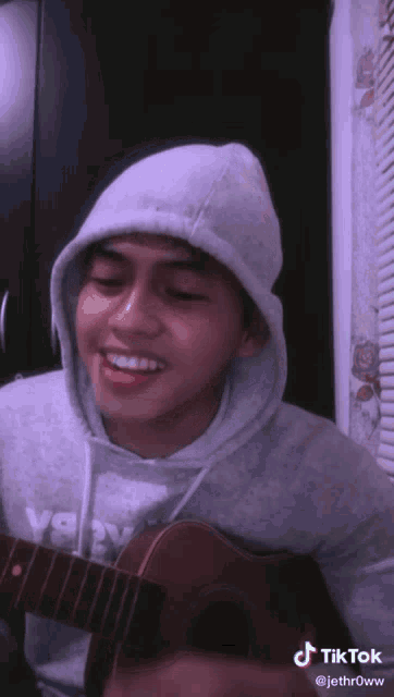 a young man wearing a grey hoodie is playing a guitar and smiling