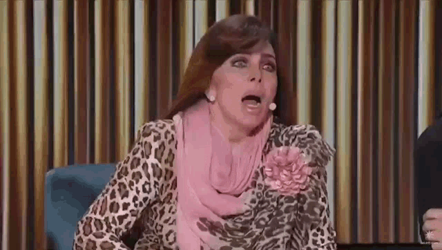 a woman wearing a leopard print dress and a pink scarf is sitting in a chair with her mouth open .