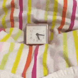 a square clock laying on a bed with a striped blanket shows the time as 4:20