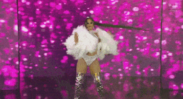 a woman in a white fur coat and silver boots is dancing in front of a pink background .
