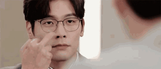 a man wearing glasses is looking at himself in the mirror and pointing at his nose .