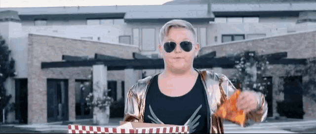 a boy wearing sunglasses and a gold jacket is holding a bag of chips in front of a large house .