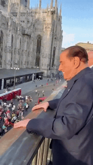 a man in a suit stands on a balcony overlooking a crowded city