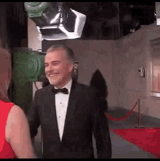a man in a tuxedo and bow tie is shaking hands with a woman in a red dress .