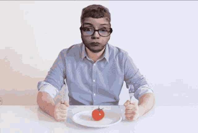 a man with glasses sits at a table with a knife and fork and a tomato on a plate