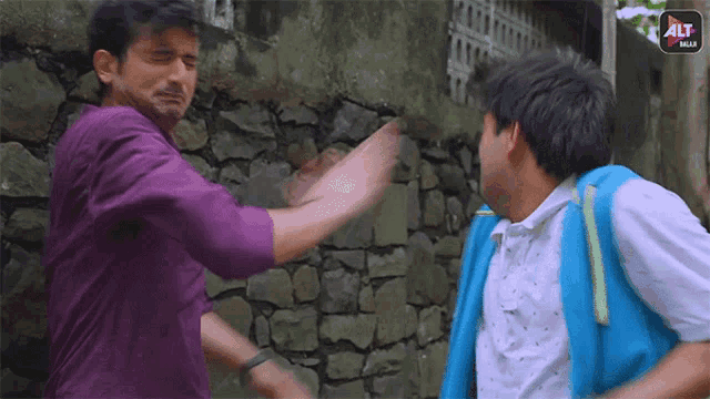 a man in a purple shirt is fighting another man in a blue jacket in front of a stone wall .