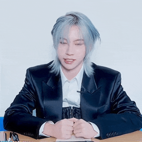 a young man with blue hair is sitting at a desk with his eyes closed
