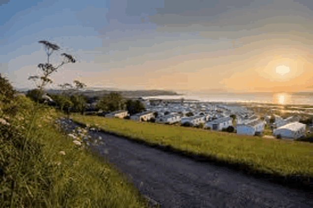 a dirt road going through a grassy field with a view of a caravan park at sunset .