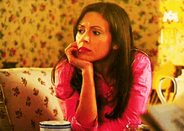 a woman in a pink shirt sits on a couch with her hand on her chin