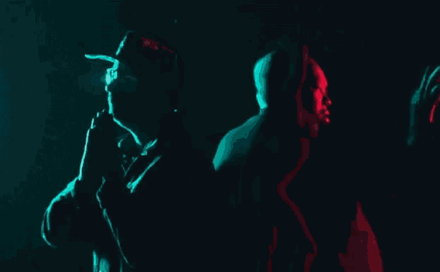 a man in a hat is standing next to another man in a hoodie in a dark room