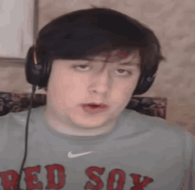a man wearing headphones and a red sox shirt looks at the camera