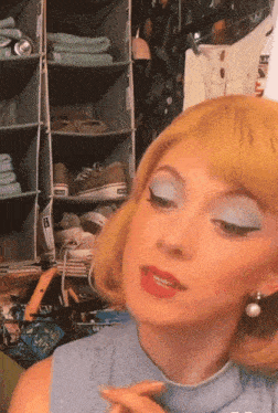 a woman wearing blue eyeshadow and pearl earrings stands in front of a closet full of shoes