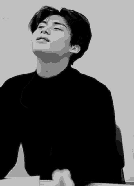 a black and white photo of a young man in a black sweater sitting at a table .