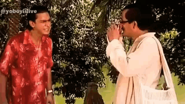 two men are standing next to each other in front of a tree and talking .