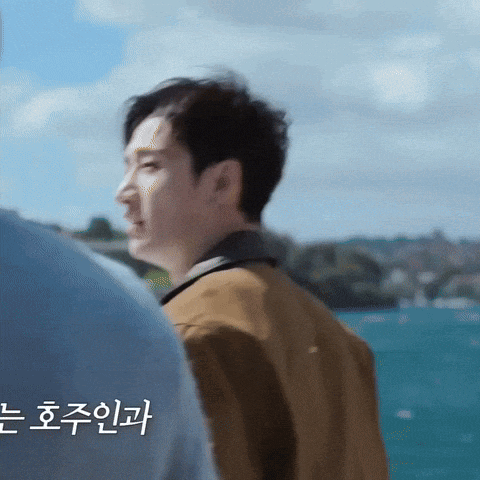 a man in a brown jacket stands in front of a body of water with korean writing on the bottom