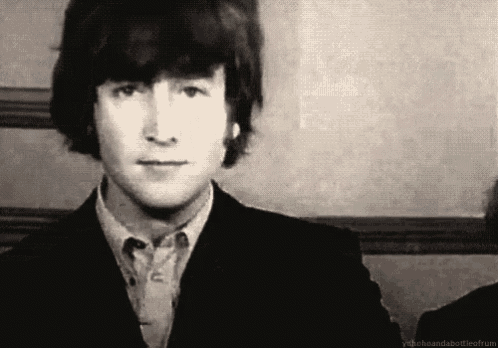a black and white photo of john lennon wearing a suit and a button down shirt .