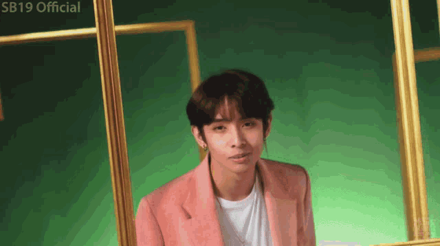 a young man in a pink jacket and white shirt stands in front of a green wall with the words sb19 official on the bottom