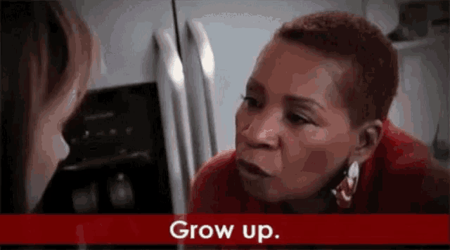 a woman in a red shirt is talking to another woman in front of a refrigerator and the words grow up are on the bottom