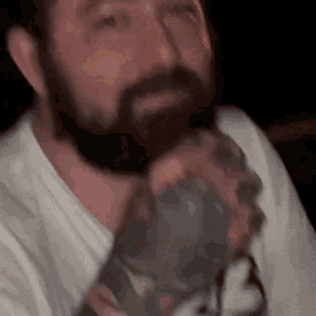 a man with a beard is drinking from a glass while wearing a white shirt .