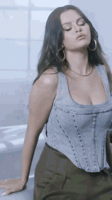 a woman wearing a denim corset top and green pants is leaning on a counter .