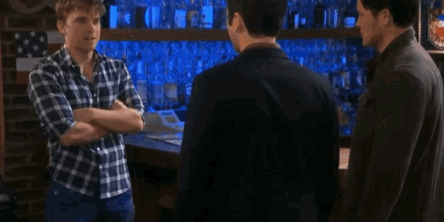 three men are standing in a bar talking to each other . one of the men is wearing a plaid shirt .