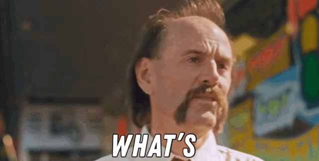 a bald man with a mustache is standing in front of a wall with the words `` what 's '' written on it .