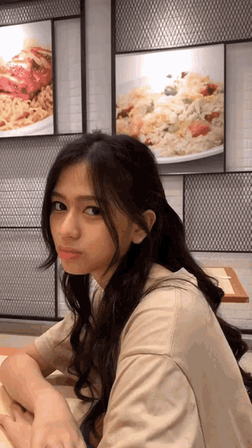 a woman sits at a table in front of a wall with pictures of food