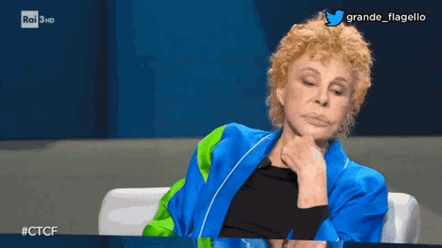 a woman in a blue jacket sits in front of a screen that says grande flagello