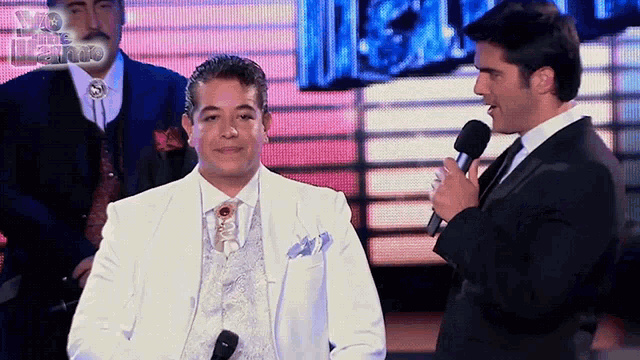 a man in a white suit stands next to another man in a black suit holding a microphone