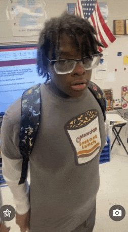a young man wearing glasses and a maruchan instant lunch t-shirt is standing in a classroom with a backpack .