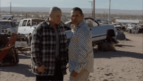 two men are standing next to each other in front of a pile of cars
