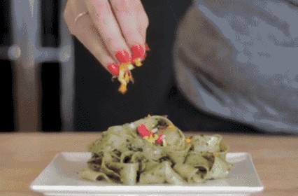 a person with red nails is adding a topping to a plate of green pasta