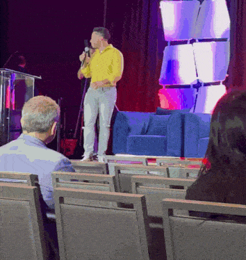 a man in a yellow shirt stands on a stage with a microphone in his hand