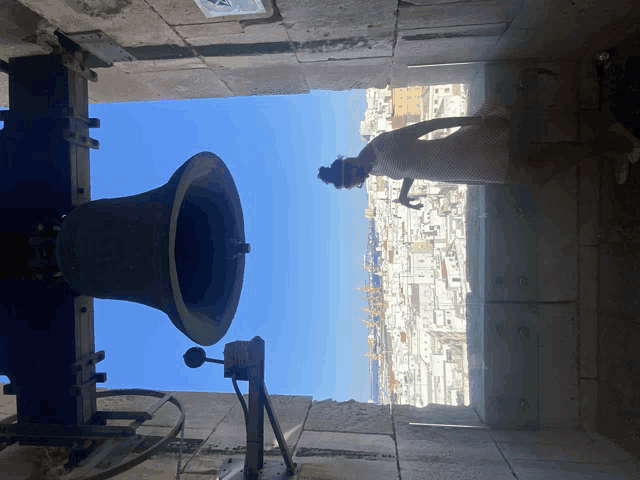 a woman stands in front of a large bell with a blue sky behind it