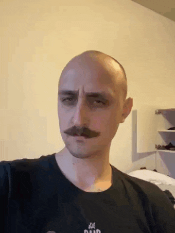 a bald man with a mustache is wearing a black shirt and looking at the camera .