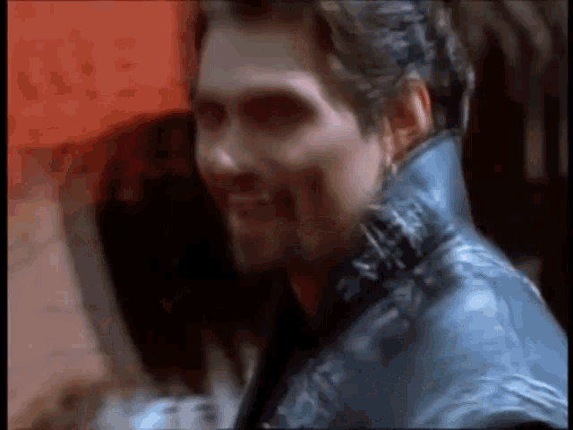 a man in a blue leather jacket is smiling and looking at the camera in a blurry photo .