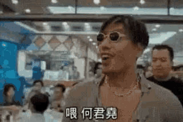 a man wearing sunglasses and a necklace is standing in a crowded room .