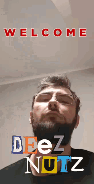 a man with glasses and a beard is standing in front of a welcome sign that says deez nuts