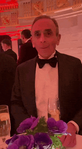 a man in a tuxedo is sitting at a table with flowers and a glass of champagne .
