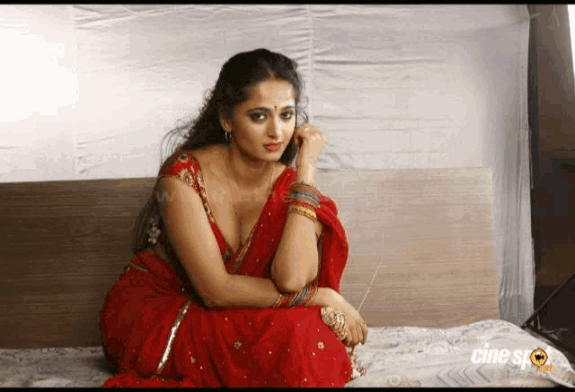 a woman in a red saree is sitting on a bed with the word cine on the bottom right