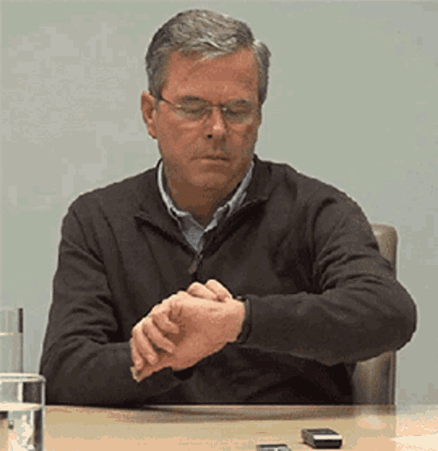 a man wearing glasses is sitting at a table and looking at his watch