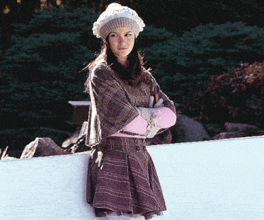a woman wearing a plaid shirt and pink mittens is leaning against a white wall