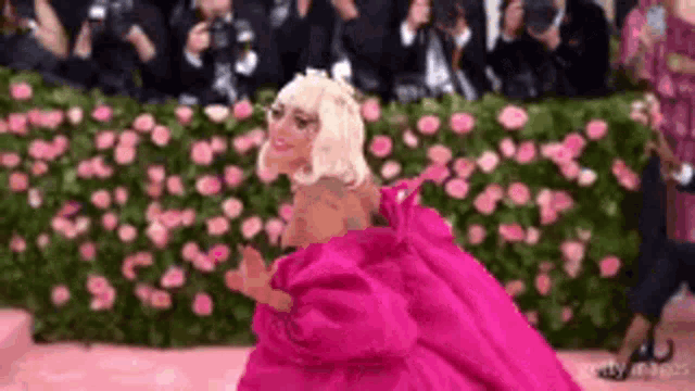 lady gaga is wearing a pink dress and standing in front of a wall of flowers .
