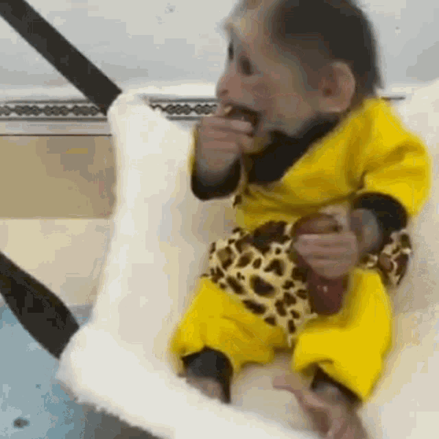 a baby monkey in a yellow outfit is sitting on a white blanket and eating a banana .