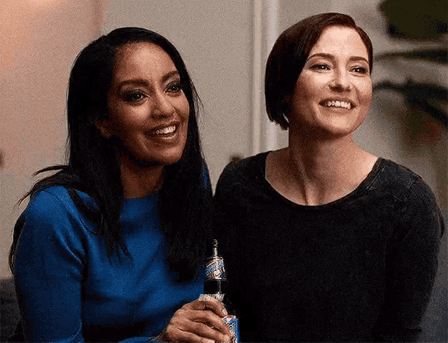 two women are sitting next to each other on a couch holding bottles of beer .