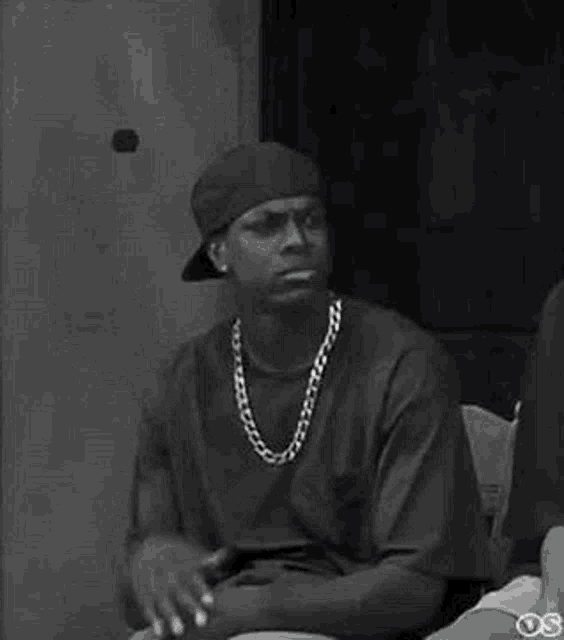a black and white photo of a man wearing a hat and necklace .