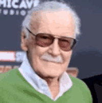 stan lee is wearing a green sweater and sunglasses and smiling .